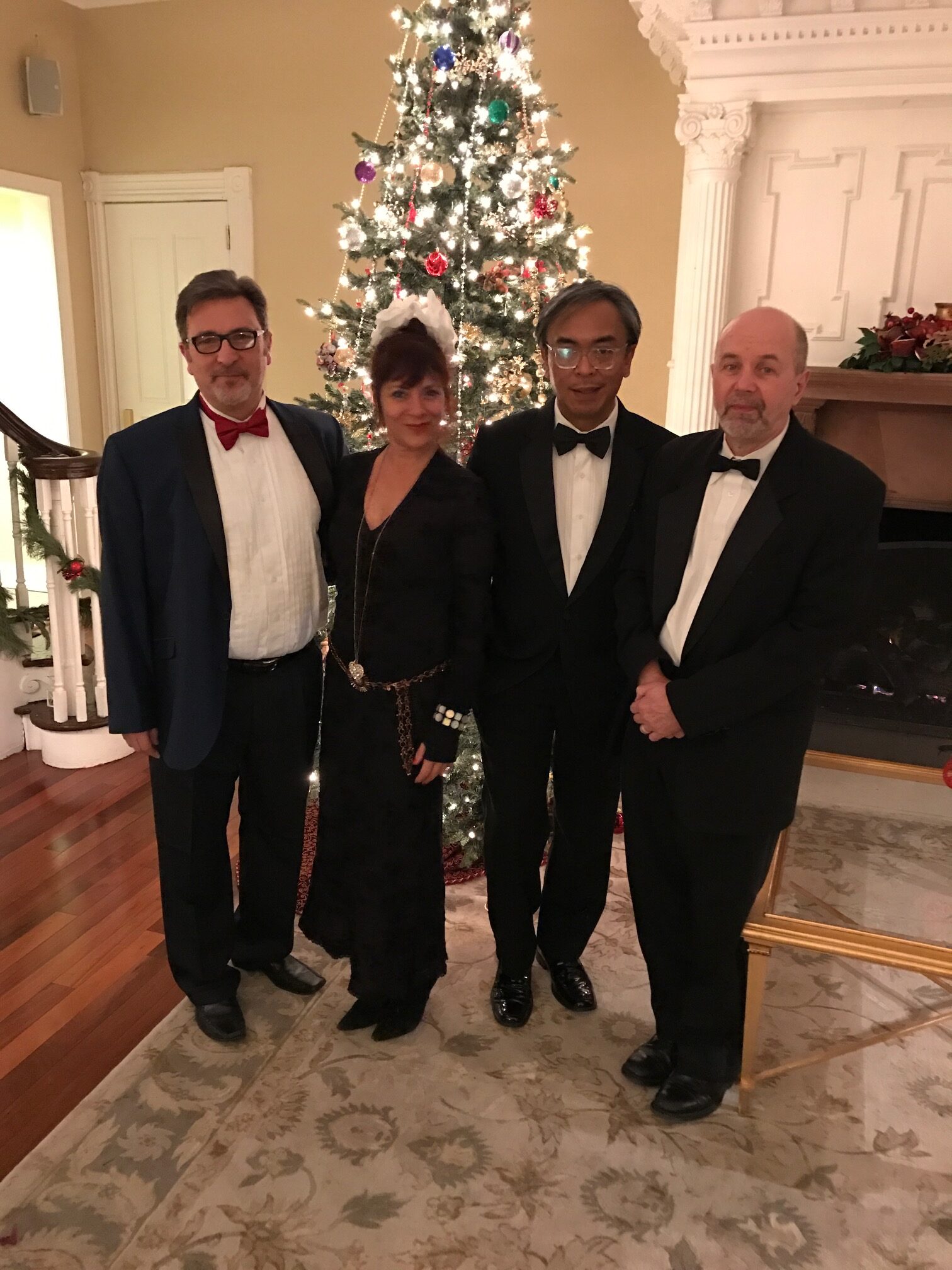 Four individuals in tuxedos stand beside a decorated Christmas tree, smiling and posing for the photo.