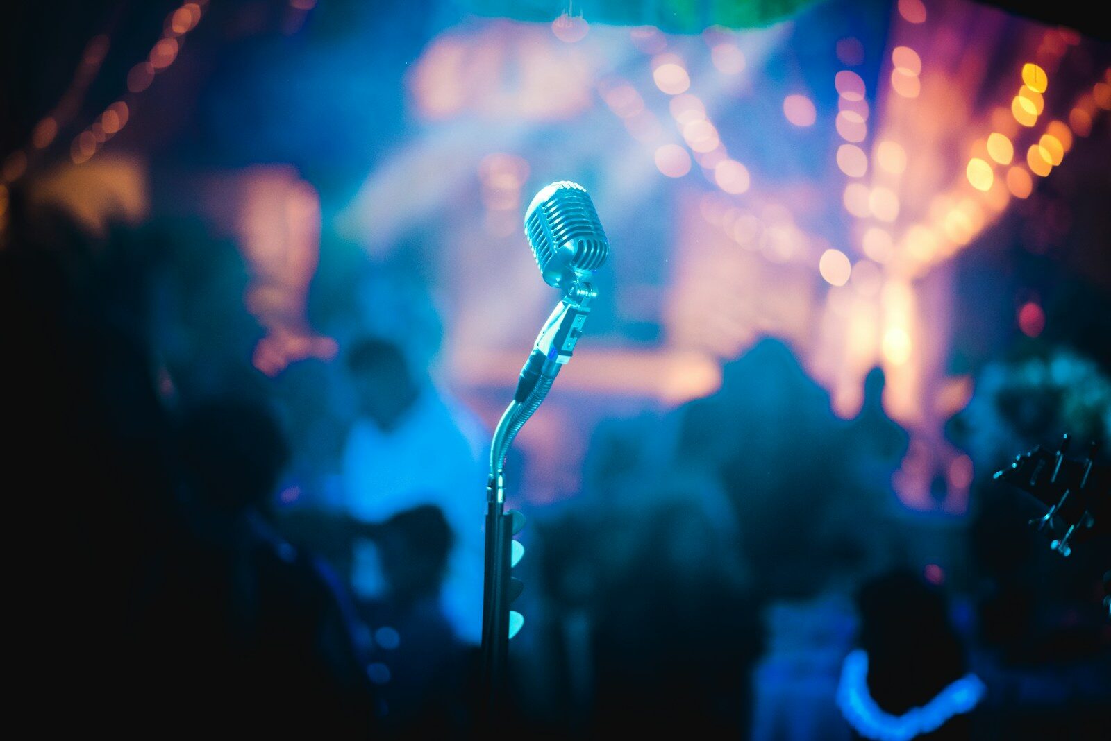 A vintage microphone stands ready on stage, surrounded by a colorful, blurred background of lights and silhouettes, capturing a lively event atmosphere.