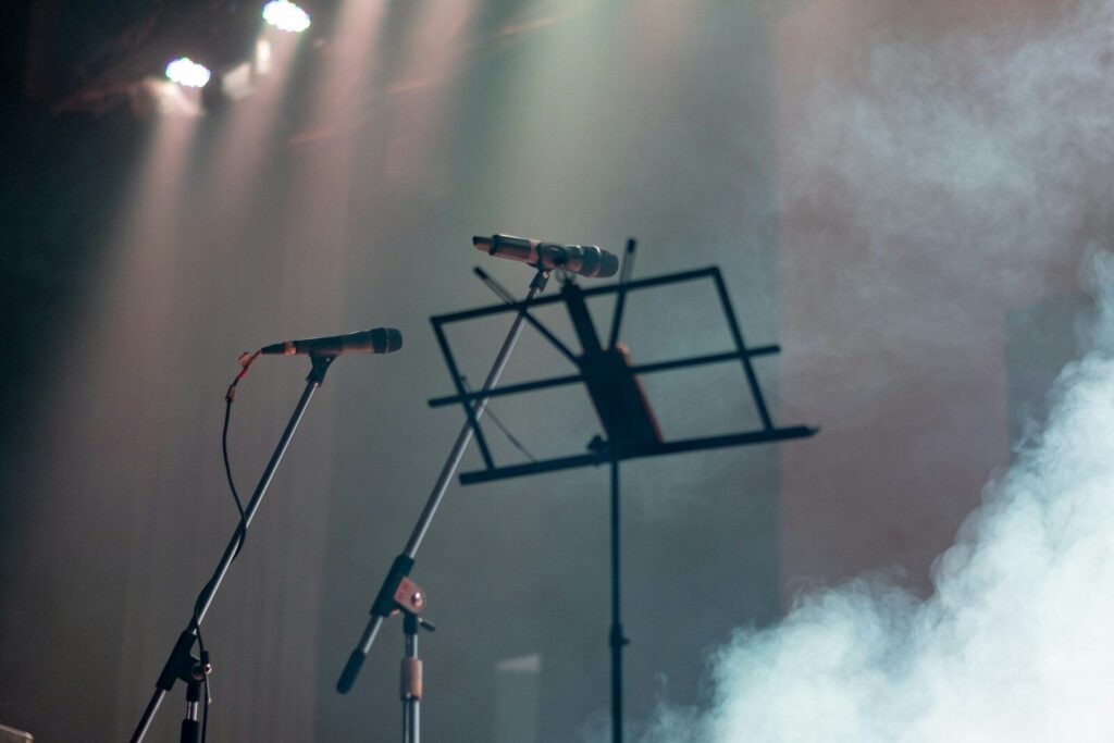 Two microphones on stands are positioned next to a music stand, surrounded by stage lights and smoke, indicating a live performance setting.